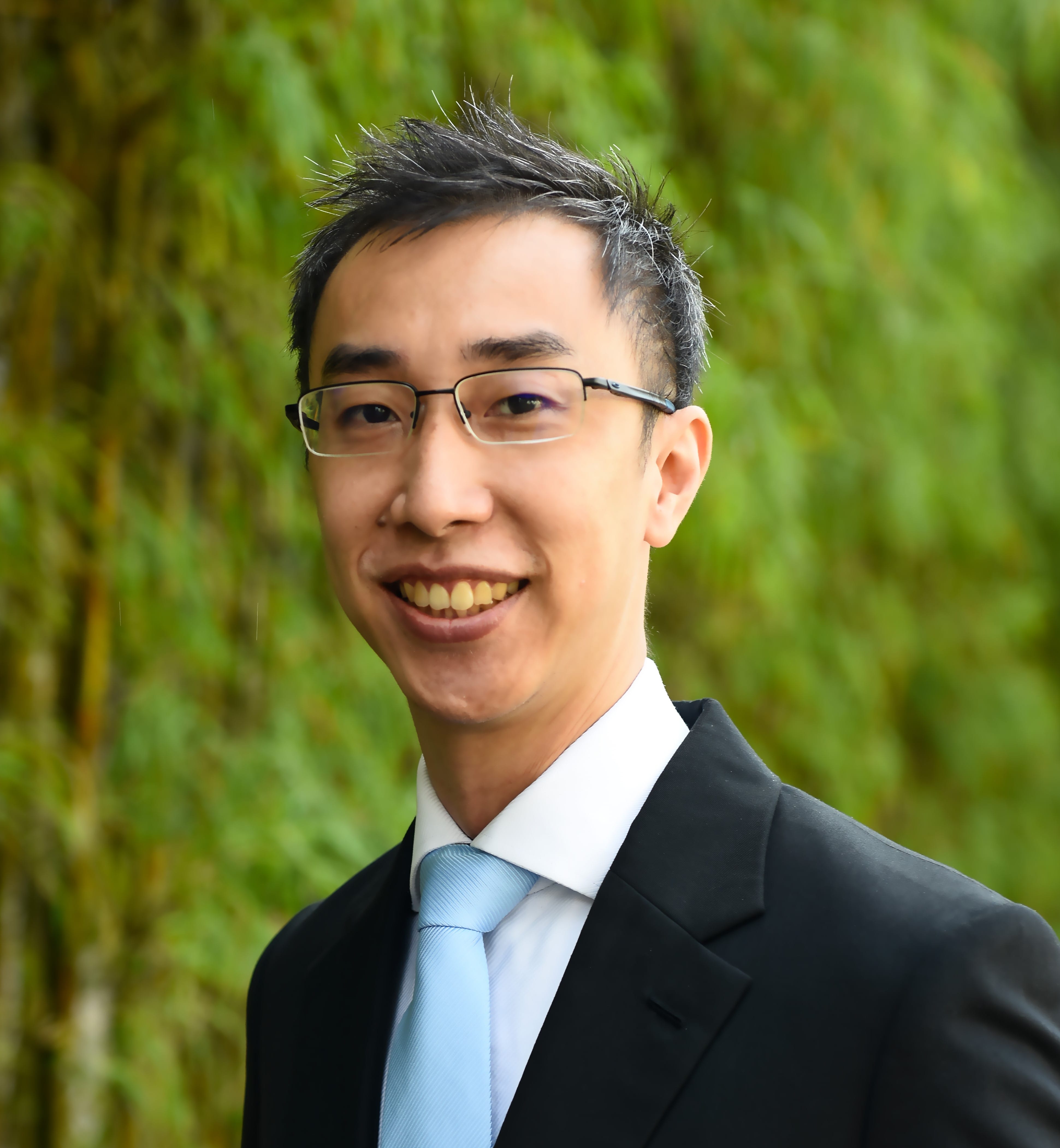 A man wearing a black suit and light blue tie stands in front of a green foliage background, smiling.